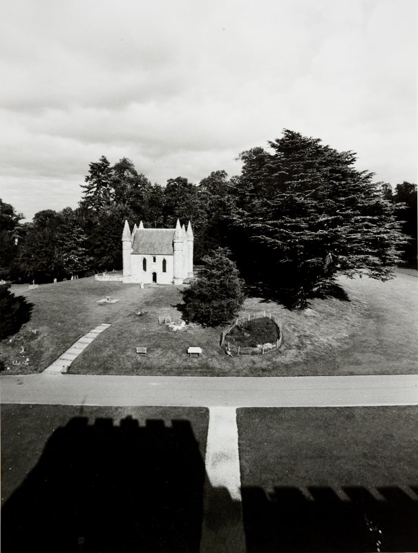 Black and white image taken from a high point of view of a white building surrounded by trees. In the forefront of the image, the dark shadow of a large wall or building in imprinted on the ground. 