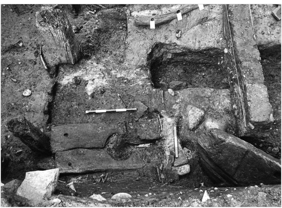 Black and white excavation photo of preserved wood and structures including a toilet seat and a wattle wall. 