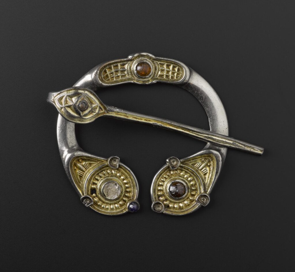 Image of a horseshoe shaped brooch with glass beads of orange, red and clear colour in the end and at the top. The brooch is silver and gold and the pin is a long silver point with a leaf shaped head, painted with the same gold leaf or yellow paint as the brooch. 