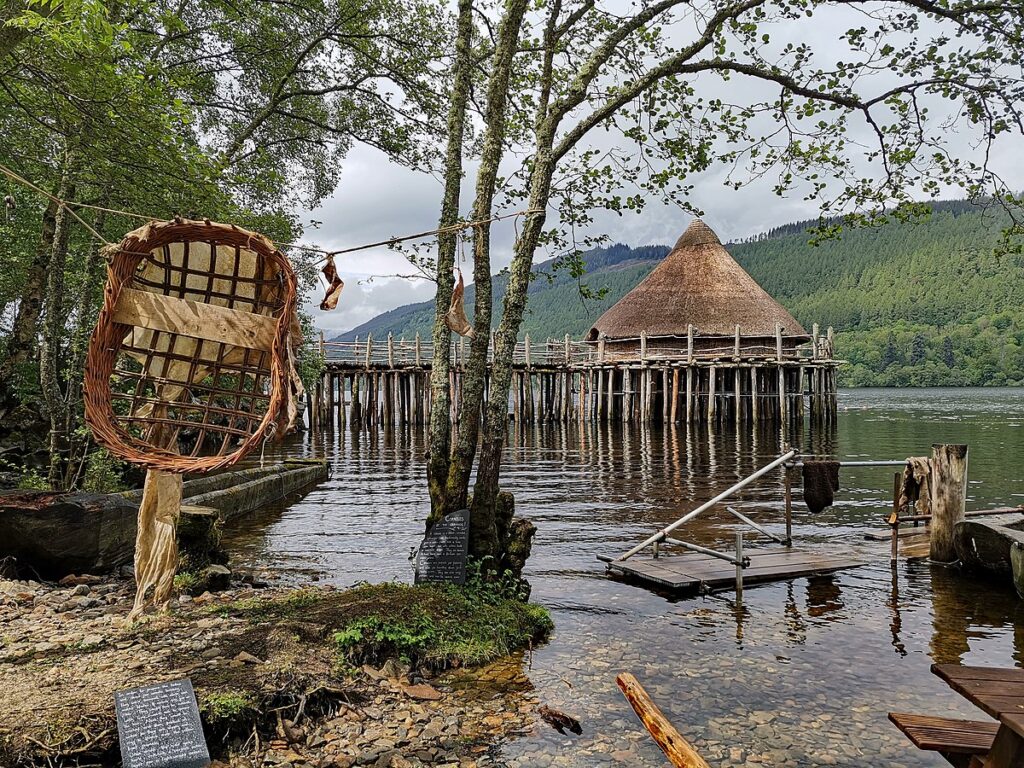 Image of a large circular building on stilts in the middle of a loch. The structure has a pointed roof and a long bridge on stilts from land. 