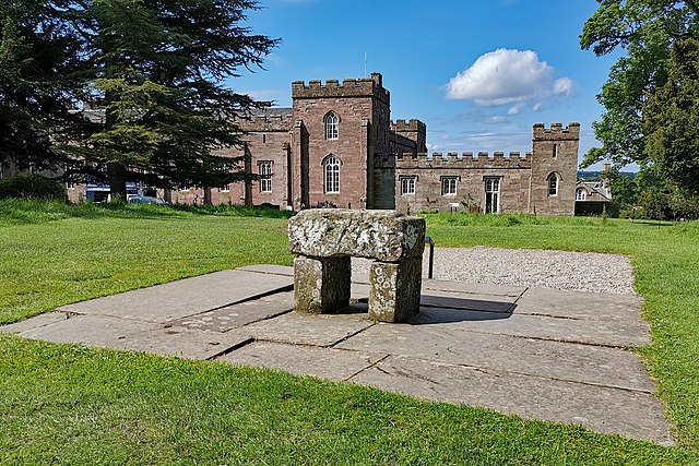 Image of a stone sculpture made of two vertical slabs holding a large rectangular stone to resemble a table or stool. The stones are placed on concrete slabs in the middle of a grassy area. A large palace can be seen in the background. 