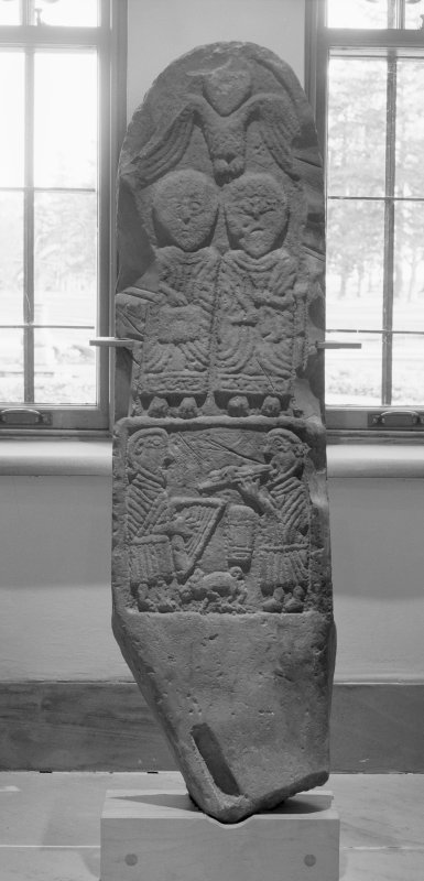 Black and white image of a long slab with carvings taken against a window. At the top is a dove, in the centre are two cherubs and at the bottom are two people playing instruments - one a harp and the other a trumpet. All carvings are done in relief. 