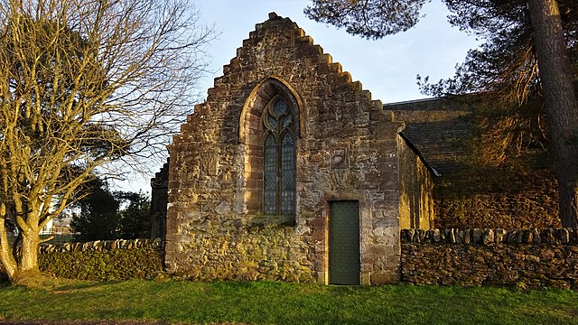 Small stone-built church with one visible doorway and a large stained glass window. The building is pointed at the top, and is adjoined to a stone wall. 