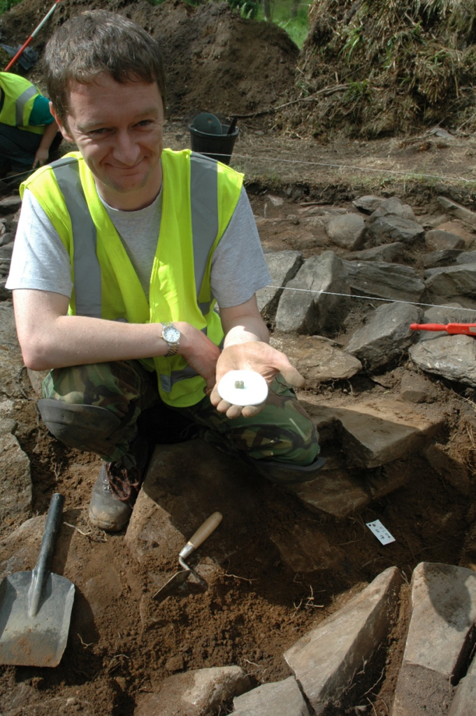 An excavator kneals in the excavation trench, holding a glass bead in one hand and leaning on his knee with the other. He is wearing a high vis vest and smiling at the camera. 