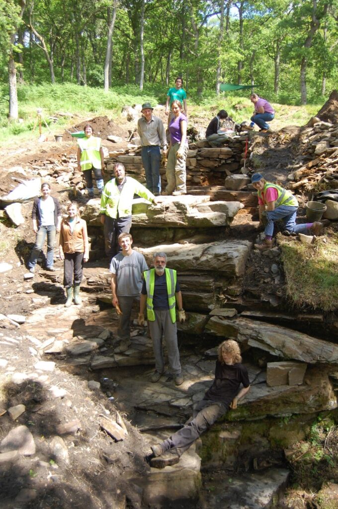 The excavation team pose for a photograph, with people standing and kneeling on five different layers of the site, showing the true scope of the Black Spout. 13 people in total are pictured wearing high vis vests and digging gear. 