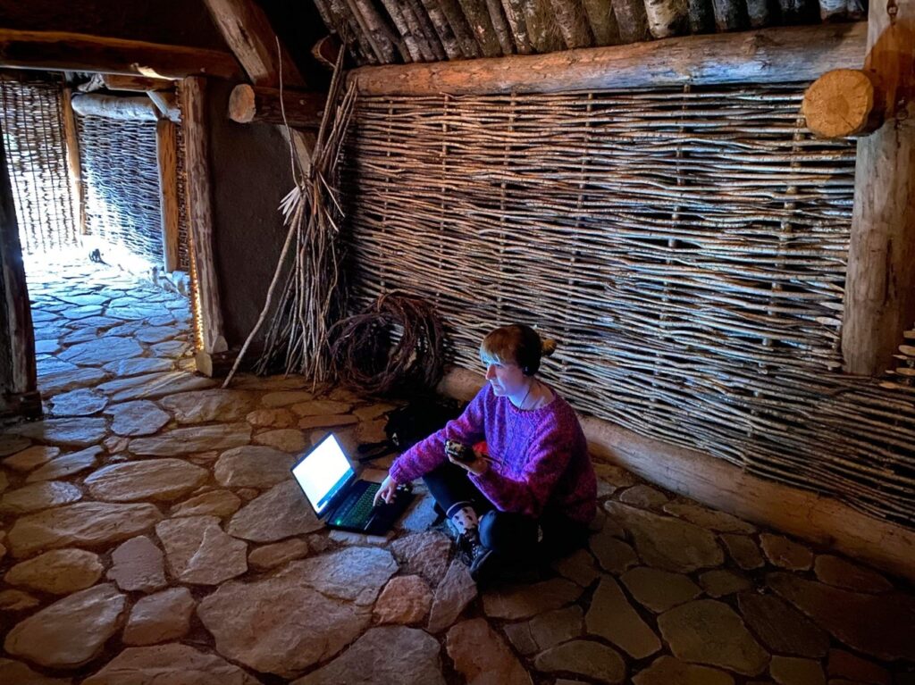 A woman wearing a purple jumper sits cross legged on the stone floor of a turf house with wattle walls behind her. She has a laptop on the ground in front of her and is holding a digital device. 