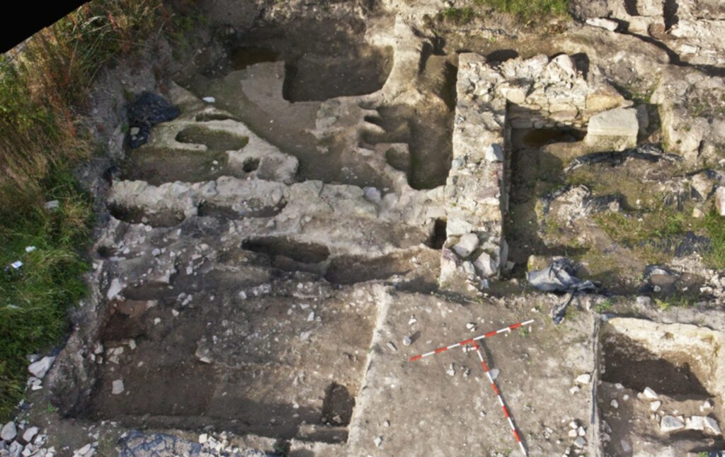 Excavation photgraph showing stonework and multiple irregular trenches. A large, thick wall separates the inner features and an external platform. 