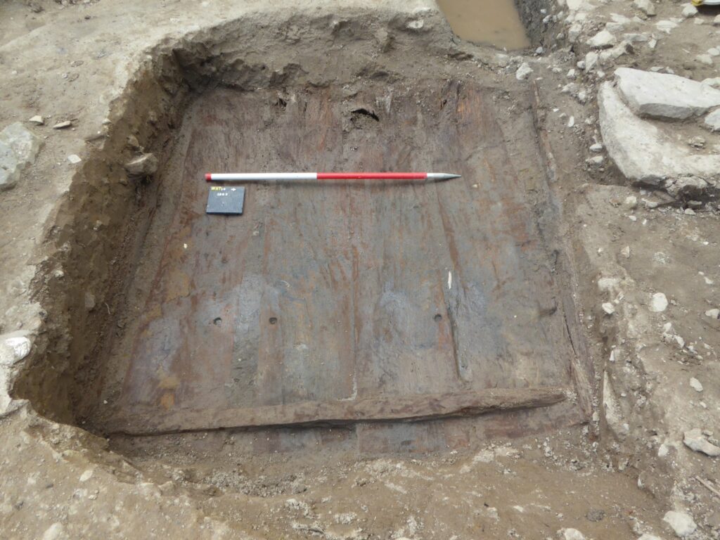 Excavation image of a flat, wooden lid to a grave. The lid is square and made of planks of wood fixed together. Some rivet holes can be seen in the wood. 