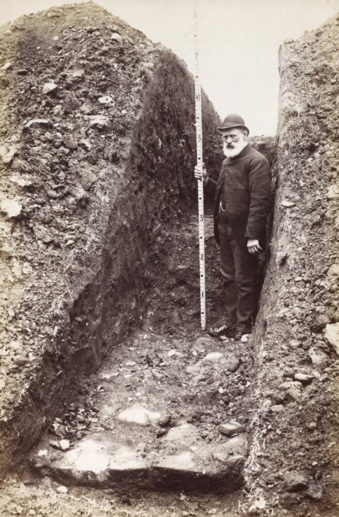 Black and white image of a man wearing a suit and bowler hat holding a metre stick. He is standing in a deep, narrow trench which looks like a hallway. 
