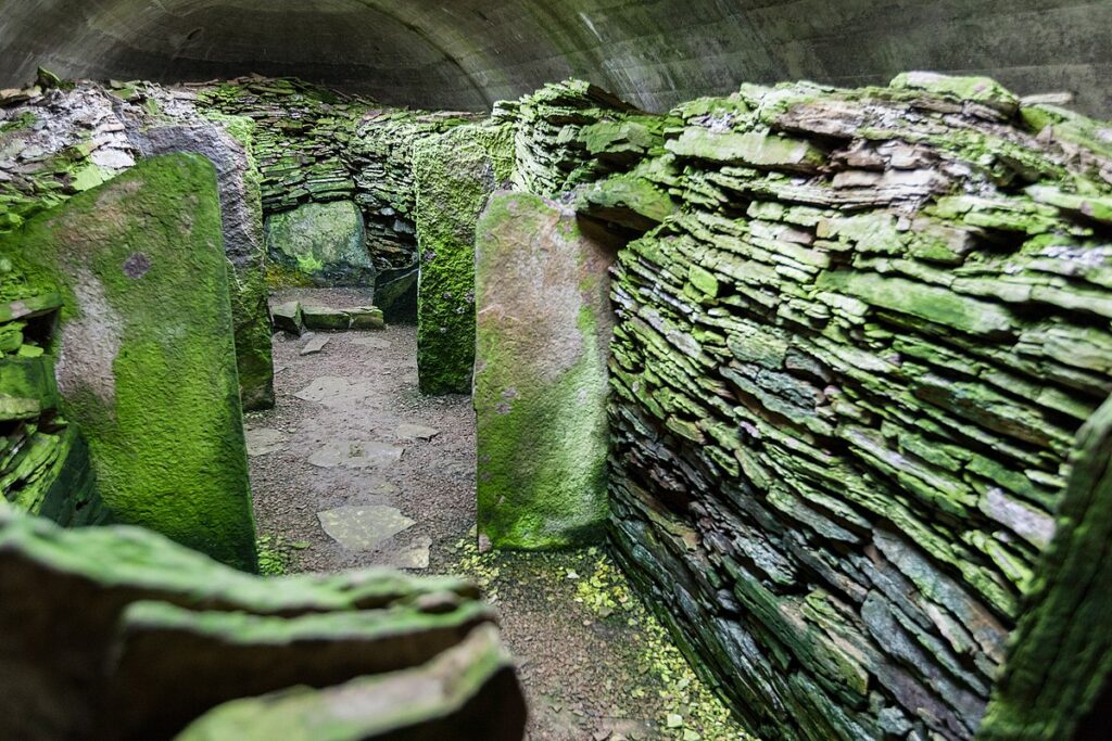 View from inside a stone-lined tomb. The walls are made with thin, grey drystone slabs withlarge upright slabs jutting out everyfew feet. All stones are covered in moss. There is a vaulted ceiling above and some dispersed slabs on the ground to create a path through the tomb. 