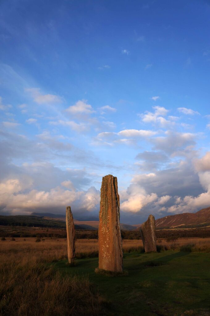 Portrait image of three of the irregular standing stones at Machrie Moor, taken at sunset with white and grey clouds on a blue sky, and an orange hue covering the stones and the landscape of hills in the background. 