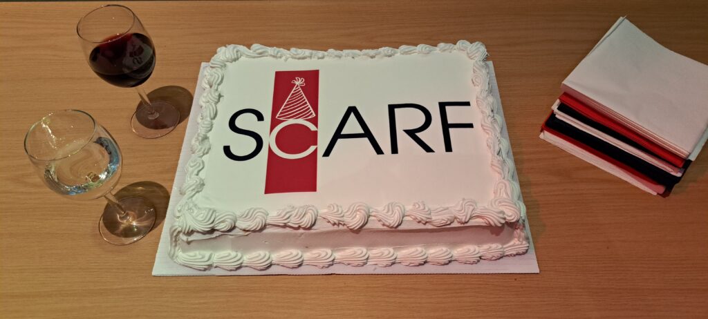 A rectangular sheet cake with glasses of white and red wine and red, blue and white napkins sits on a table. The cake has the ScARF logo with a bithday hat above the 'C'. 