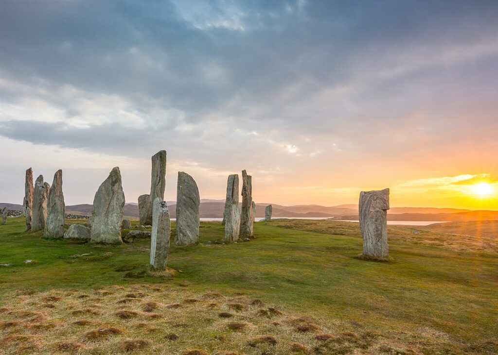 A cluster of pointed and sub-rectangular standing stones sits to the left of the image on a grass covered hill with water in the background. To the right is a single standing stone, rectangular in shape. The far right shows the sun setting, with orange light flairs coming from it. 