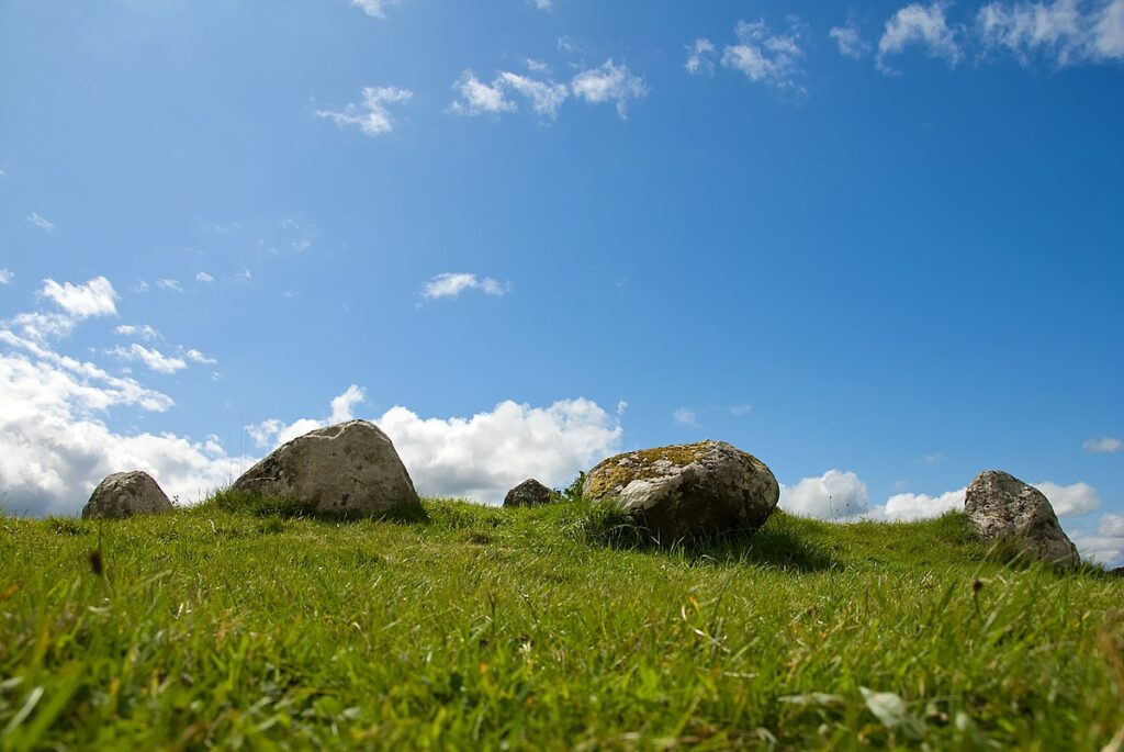 Eye-level photo of rounded stones from outside a stone circle. The camera is at grass-level, with green grass out of focus in the foreground. The sky is blue with with clouds. 