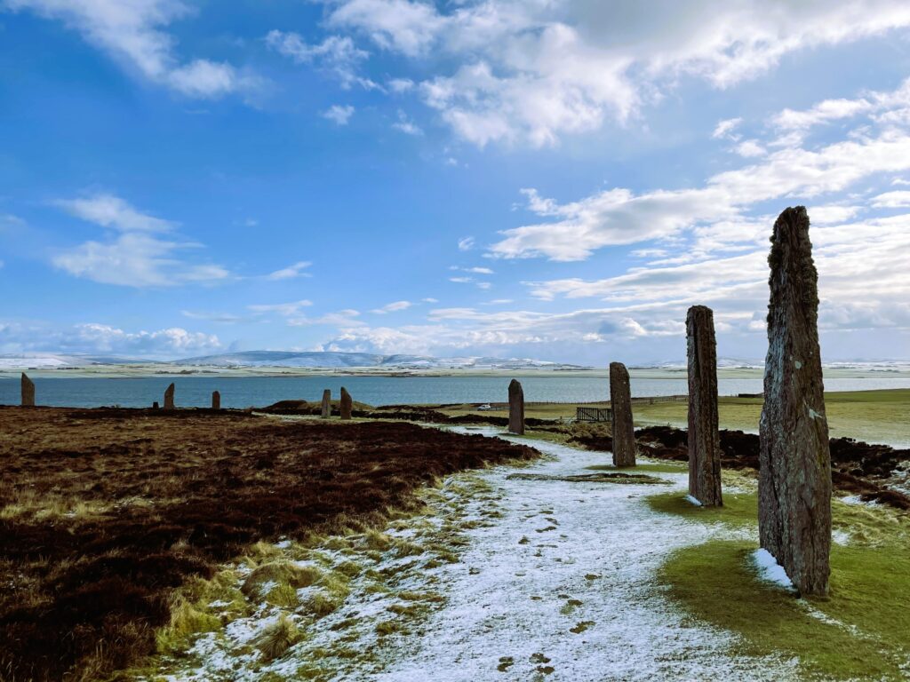 Photograph taken from inside a stone circle on a snowy, but sunny day. Nine standing stones, all sub-rectangular, are seen, becoming smaller to the left of the image. The sea can be seen in the background. 