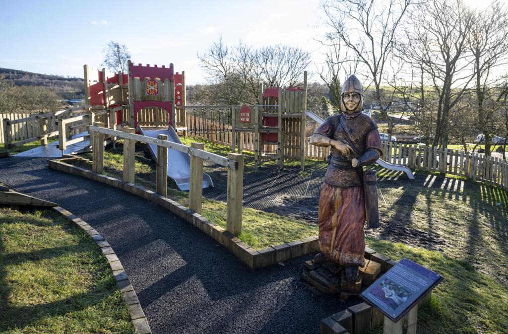 A playpark with a Roman soldier statue standing at the gate, red and yellow fort wall details on the slides and climbing frame, and enclosed in a wooden fence. 