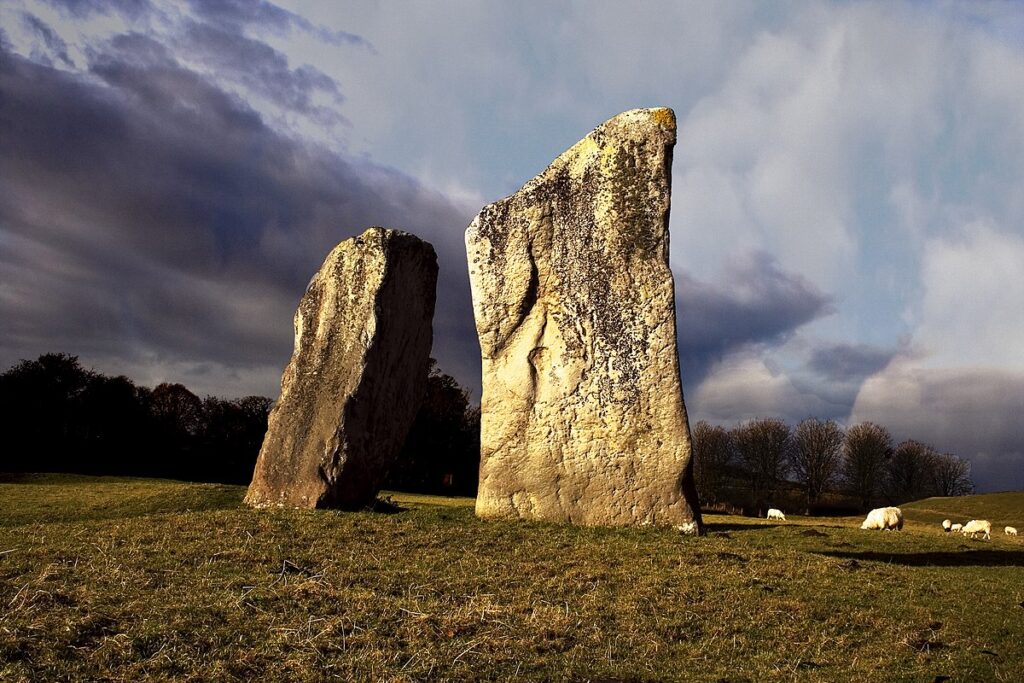 Two large standing stones sit on a grassy field with a dramatic, dark blue, grey and black sky behind them. Sheep can be seen in the distance grazing. 