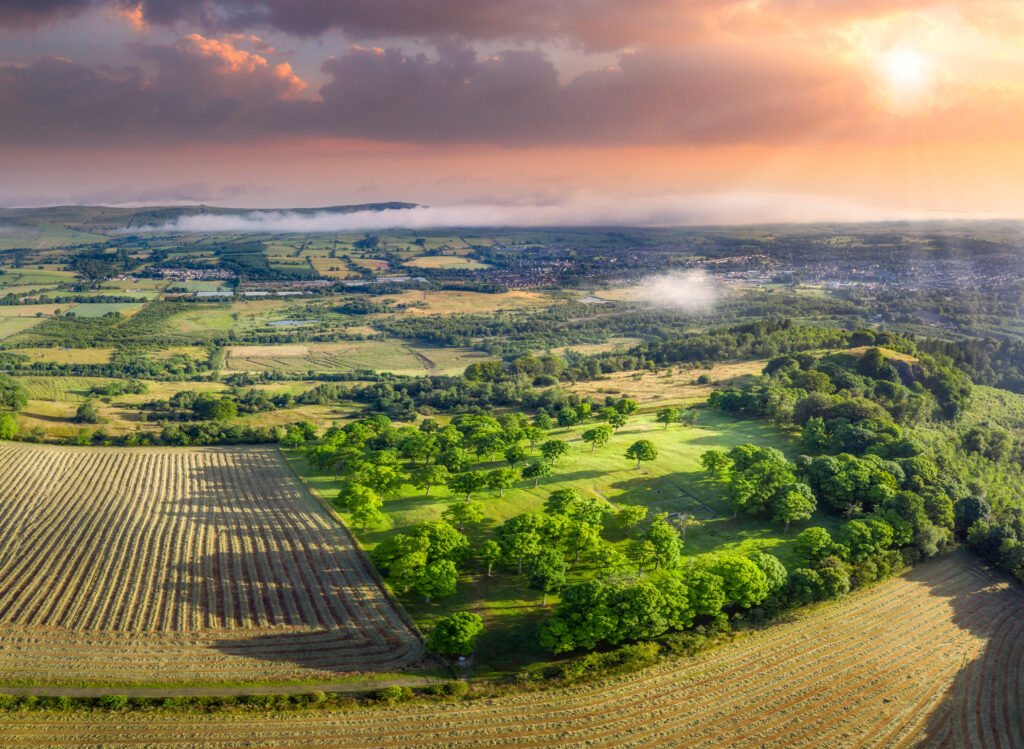 Atmospheric landscape shot of a of fields and forests from an oblique aerial view. The sky is red and orange, the sun it peering through the clouds. A rectangular cropmark can be seen on the left side in a field. 