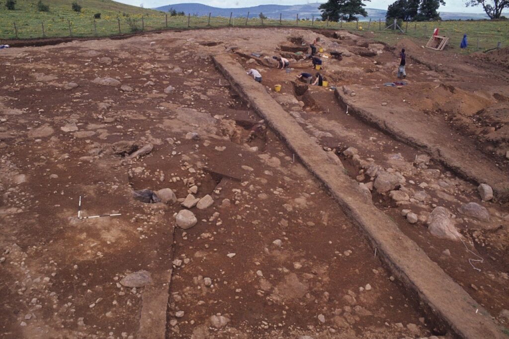 A colour photograph of an excavation with brown soil, linear stone stuctures and people excavating towards the back of the image.