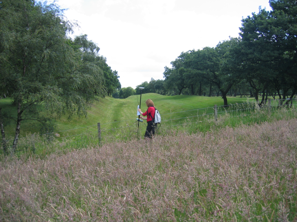 A woman wearing a red long sleeve top and a white backpack walks through  heather-filled field holding a surveying rod which is a foot taller than her. 