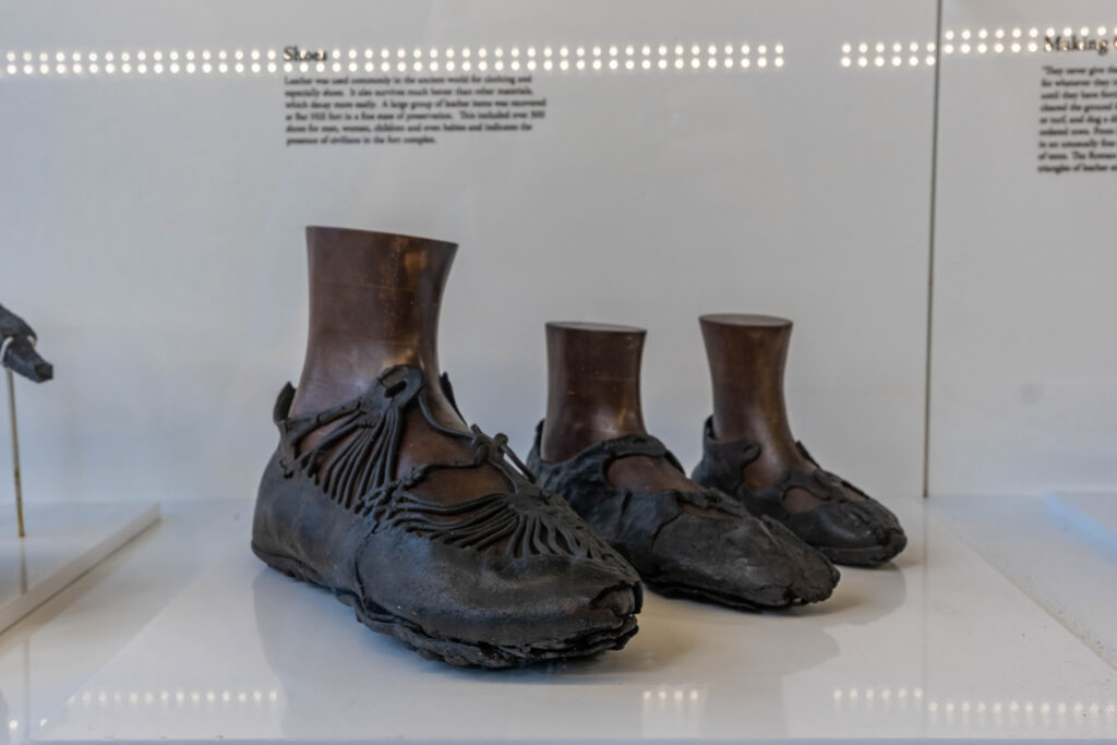 Museum image of three leather shoes in lessening size place in a row inside a glass case. The shoes are all placed on foot shaped stands and are black, detailed leather with loops for laces. 