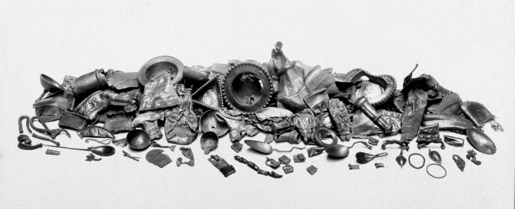 An arranged assemblage of the Traprain treasure in a long, forward facing row with some items spread to the front and the larger items piled together in a row at the back. Jewellery, small metal objects and cutlery make up the hoard. The image is in black and white with a white background. 
