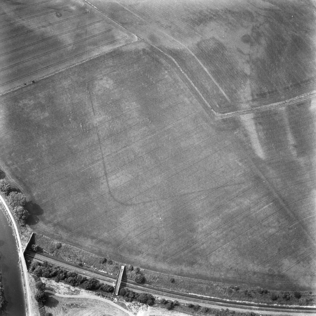 Black and white aerial photograph of a field with a curved edged rectangular cropmark near a modern road and river. 