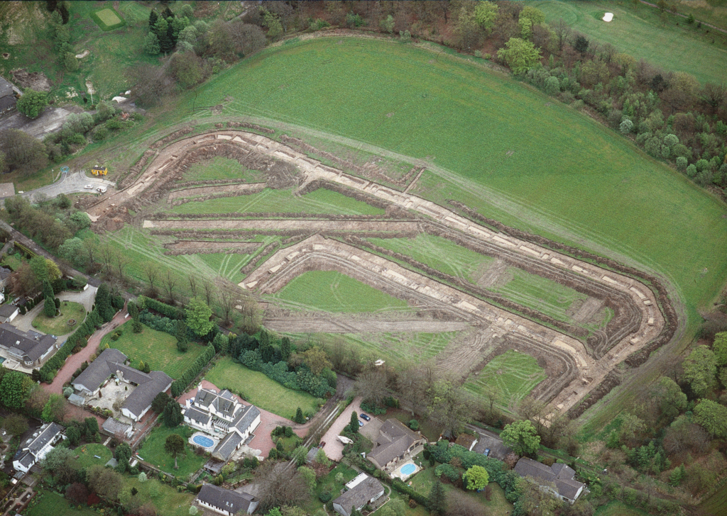 Oblique aerial view of trenches showing the exact layout of the ditches and walls of a rectangular fort. The outer rectangle takes up most of the grass-covered area. Houses are seen in the foreground. 