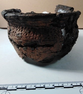 Dark brown-black bowl-shaped vessel with a faint checked pattern around the rim, with a dot in each square. It appears broken and pieced back together. 