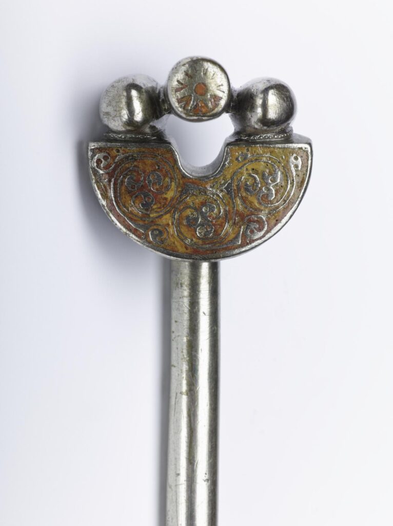 Closeup against a white background of a shiny silver handpin, with a long straight pin and a U-shaped head, with orange-red details and solid silver ball decorations. The object is slightly scratched but shiny and in good condition. 
