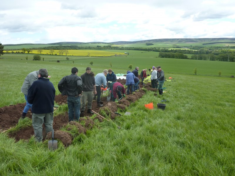 A long, narrow trench stretches horizontally across the image. Upwards of 15 people stand around or in the trench, some with shovels. The surrounding landscape is green grass, trees and a yellow rapeseed farm to the top left of the image. 