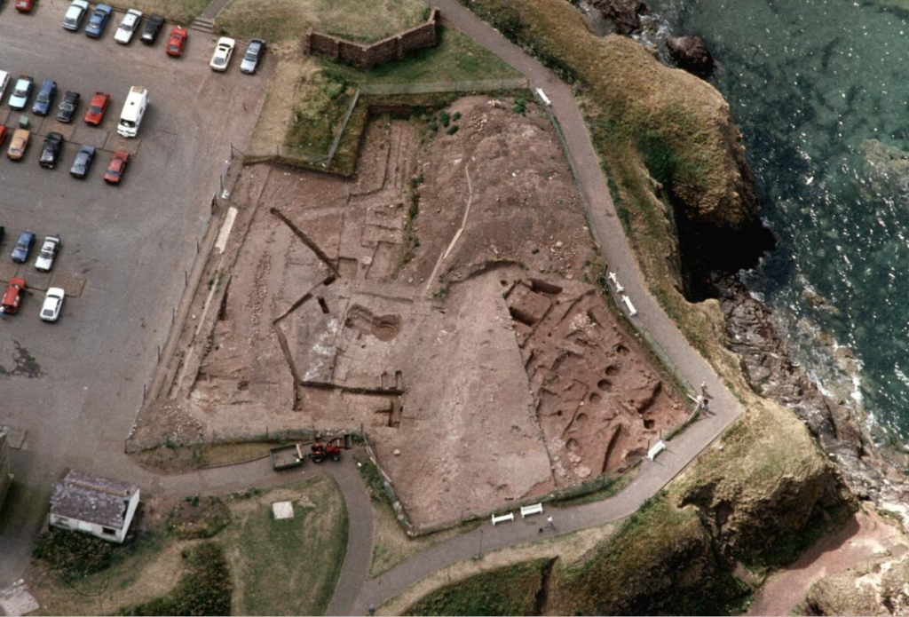 Aerial photograph of an excavation site. on the left is a car park, and on the right is a cliff overlooking water.