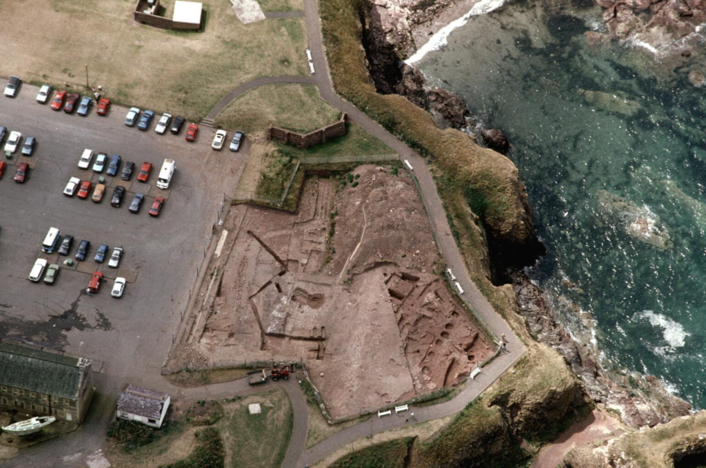 Aerial photograph of an excavation site. on the left is a car park, and on the right is a cliff overlooking water.