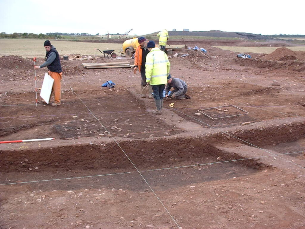 Photograph of people working at an excavation site. The ground is divided into quadrants by rope, and three of the people are gathered around one of these quadrants.