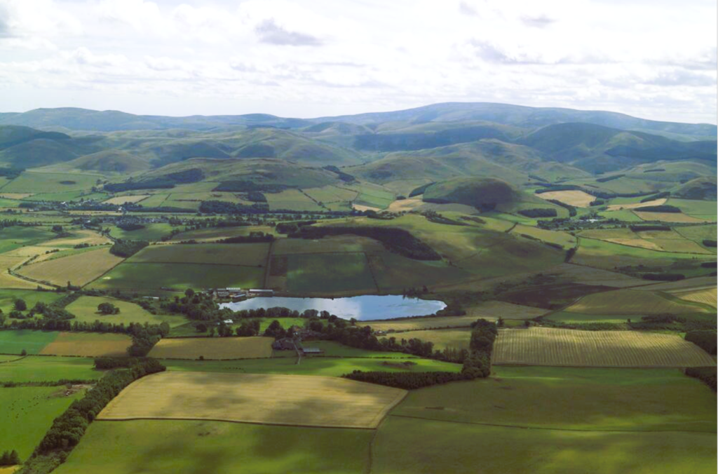 Aerial landscape photograph of hilly regions. There is a loch in the foreground, an remains of the forts in the distance.