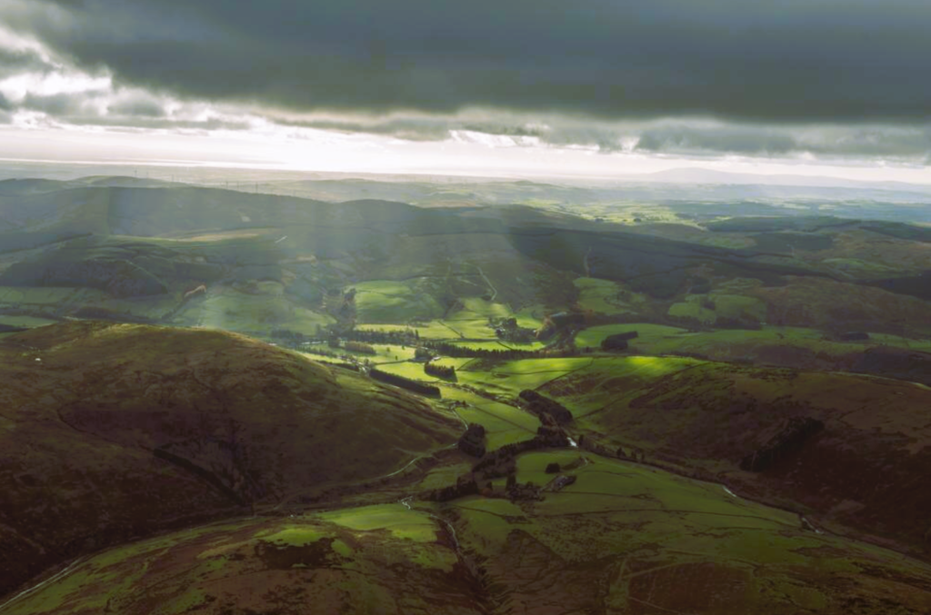 Aerial view of the hilly landscape. The view is dark, with rays of sunshine peaking through the clouds