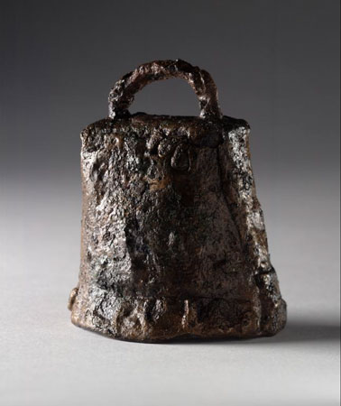 Image of a sub-rectangular rusted, black and brown iron bell against a grey background. The semi-circular handle across the top is miss-shaped and the corrosion has left an uneven surface across the whole bell. 