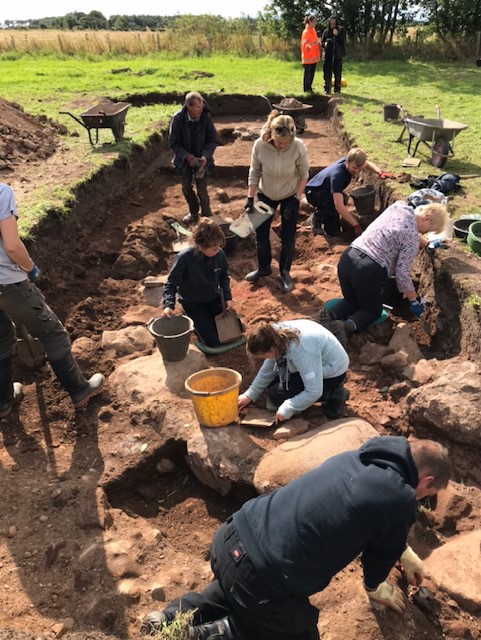Photograph of a group of 10 people taking part in an excavation. The picture is lively, with people crouched, standing and digging in the dirt.