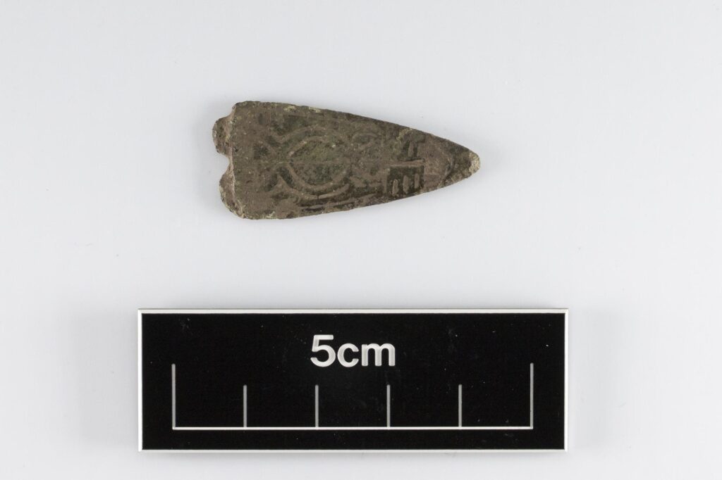 Close-up of a triangular strap end with an engraved pattern. There is a ruler indicating that it is about 15cm in size
