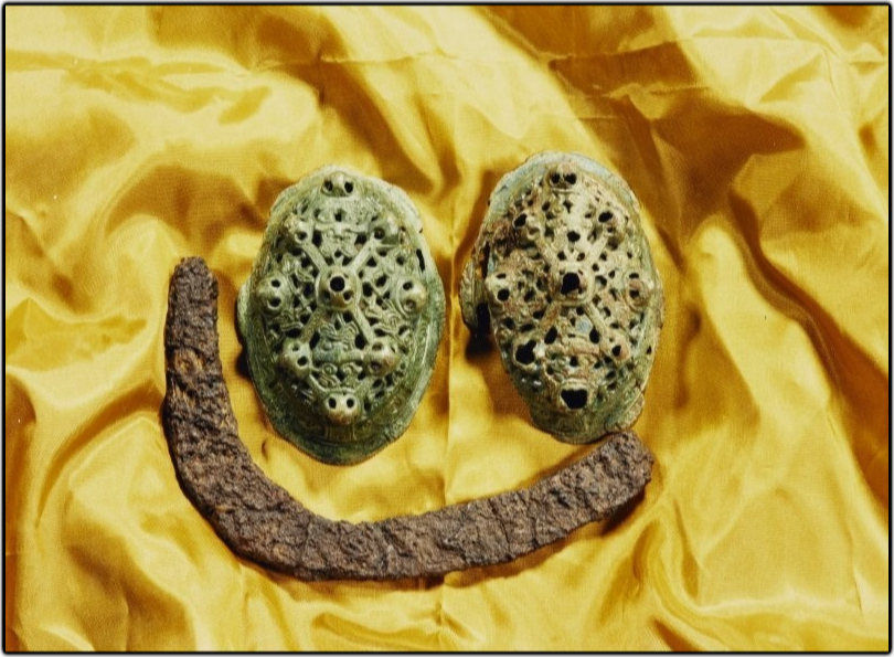 Two oval, green-coloured brooches, with a latticed pattern, and a corroded U-shaped sickle lie on a yellow silky material in the shape of a smiley face. 
