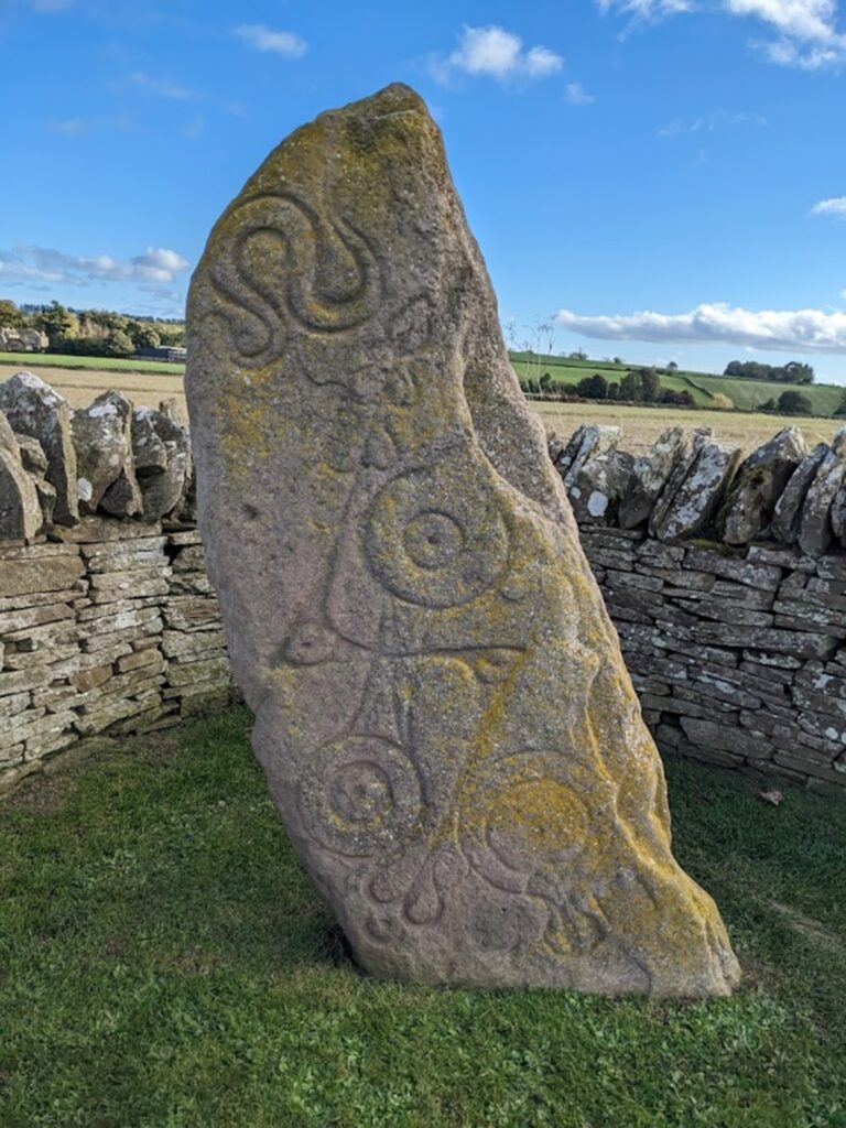 Image of a crescent-shaped standing stone within a drystone enclosure. The stone is decorated with carvings including a z-rod, serpent, mirror and doudle disk. 