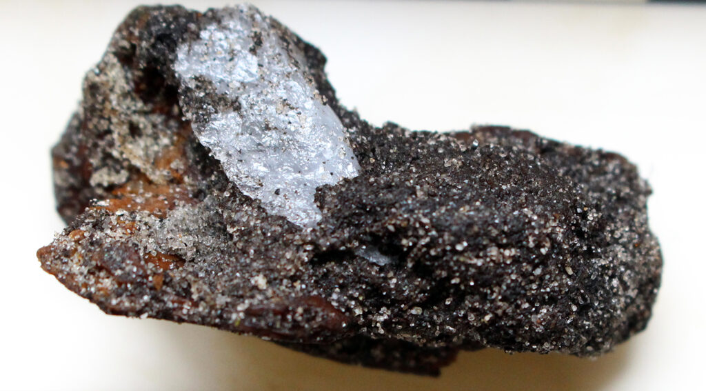 Closeup image of a black abd grey piece of quartz stone against a white background. One part of the stone is flat and shiny and the rest appears rough, like the texture of sand. 