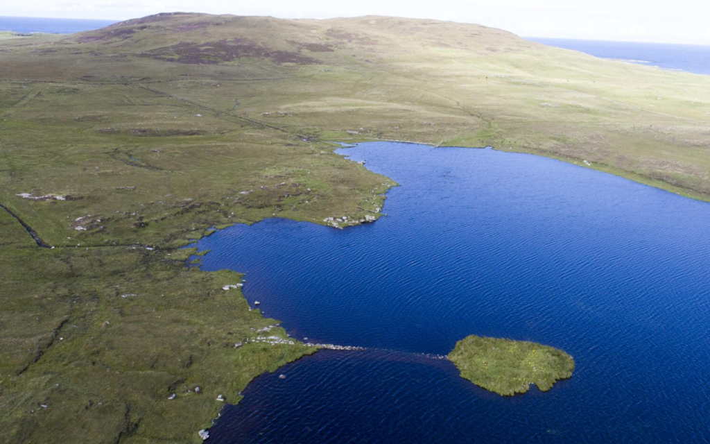 Oblique aerial view of a small island in the middle of a large, blue, lbody of water. The mainland is gree and vast. The island and the mainland seem to be connected by a very narrow and faint path, likely made of stones and sand.
