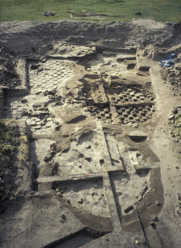 Oblique aerial view of a colpex open excavation trench with two roundhouses and the surrounding area being excavated to various depths using a grid system. 