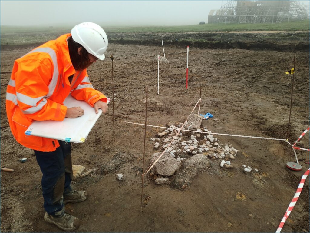 Photograph of a person wearing an orange high-vis jacket using a white drawing board to sketch a small pile of small, white pebbles, with four larger, grey boulders on the far and near side of the pebbles. In the background, a modern building can be seen under construction, with scaffolding completely surrounding the building. 