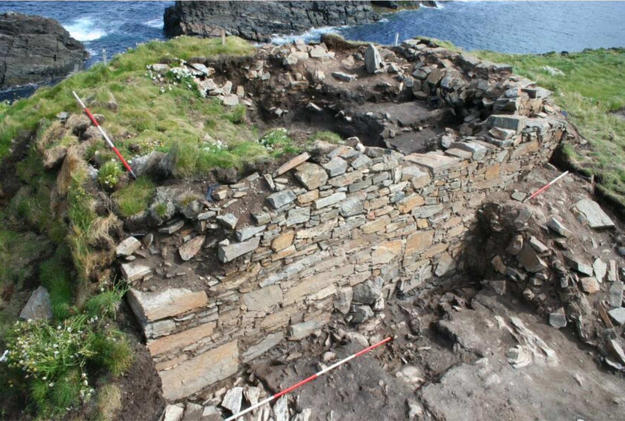 Image of a stone wall with measuring rods placed to show the size. Grass is growing over the top of the ruined structure and a body of water can be seen in the background.