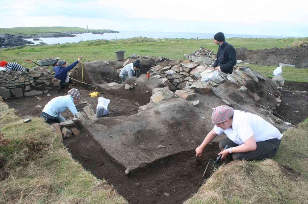 Six people in a trench excavation a stone structure. Some are trowelling while other measure features and draw plans.