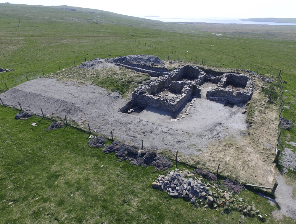 Oblique aerial view of a ruined grey stone structure. The structures is made up of walls in and L shape, with square corners. The structure appears to be on a grey platforn which is surrounded by a wire fence. The fence is surrounded by a vast, grassy field. 