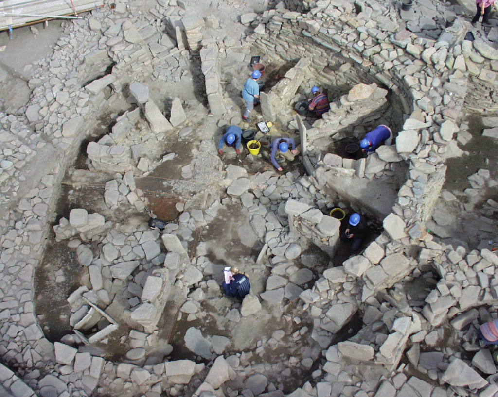 Aerial view of an active excavations in a complex stone structure with a thick, round outer wall and straight internal walls which resemble the spokes of a wheel. 10 people can be seen crouching and standing inside the structure, taking measurements and drawing using clipboards.