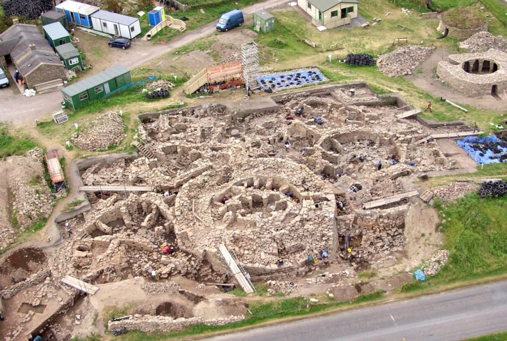 Oblique aerial view of a large open excavation site taken from a distance to show the full site. Multiple rounded stone buildings are built tightly into the open area, with the largest structure in the centre.
